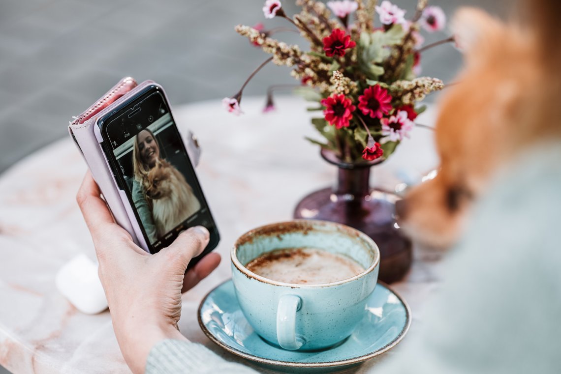 En person tar selvportrett av seg selv og hunden hvor blomster og en kaffekopp er synlig i bakgrunnen